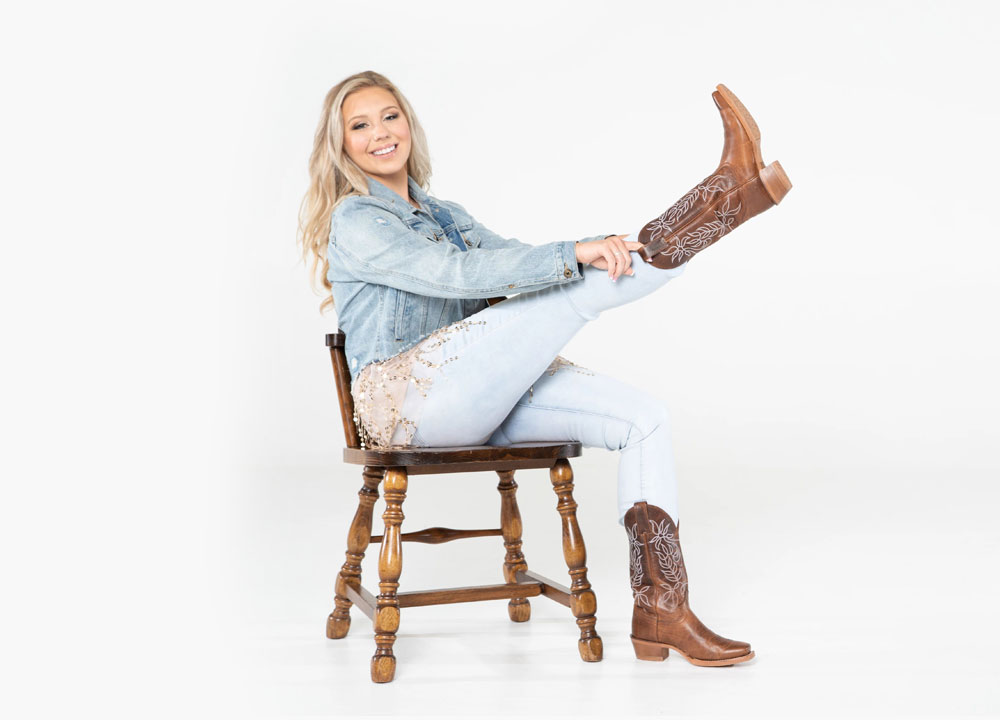 A woman wearing a jean jacket and jeans, sitting in a chair putting on cowboy boots.